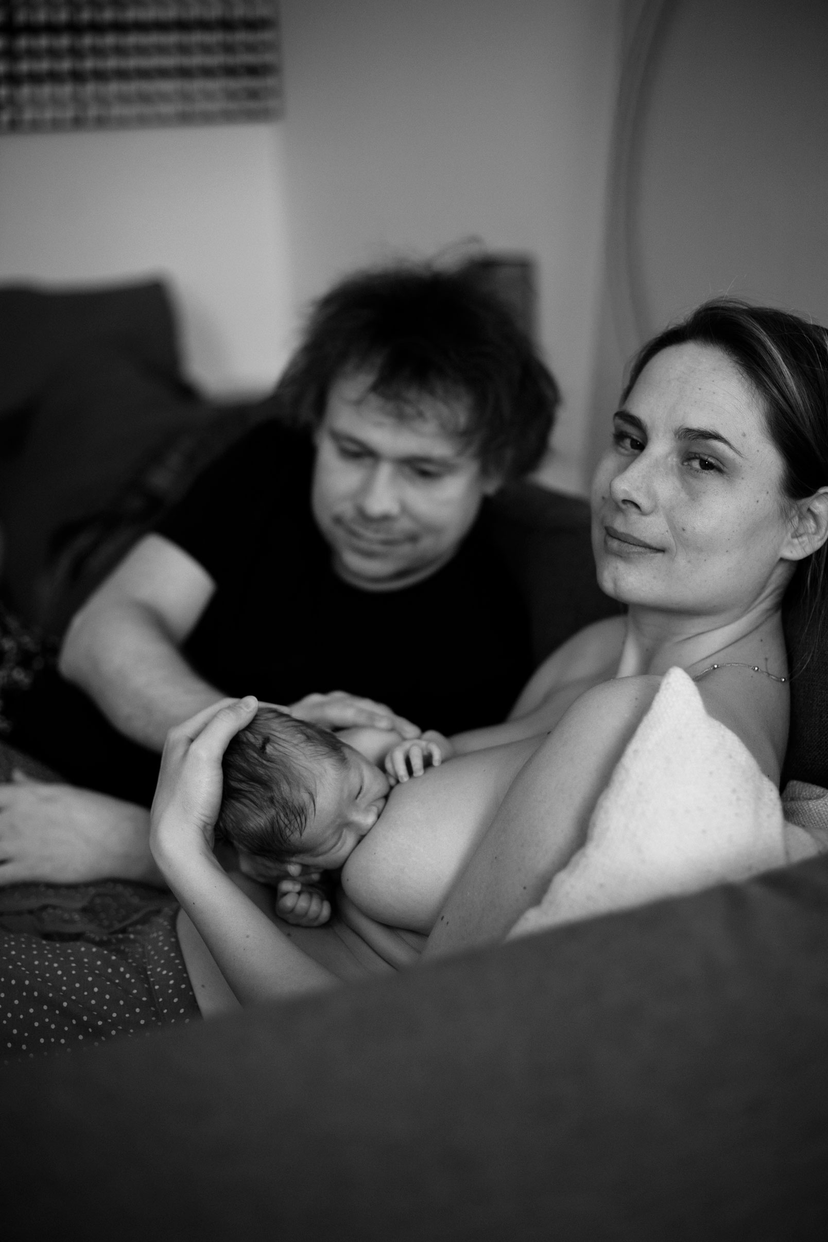 Neugeborenenfotos Berlin - Müde glückliche Mama beim Stillen mit Papa im Hintergrund